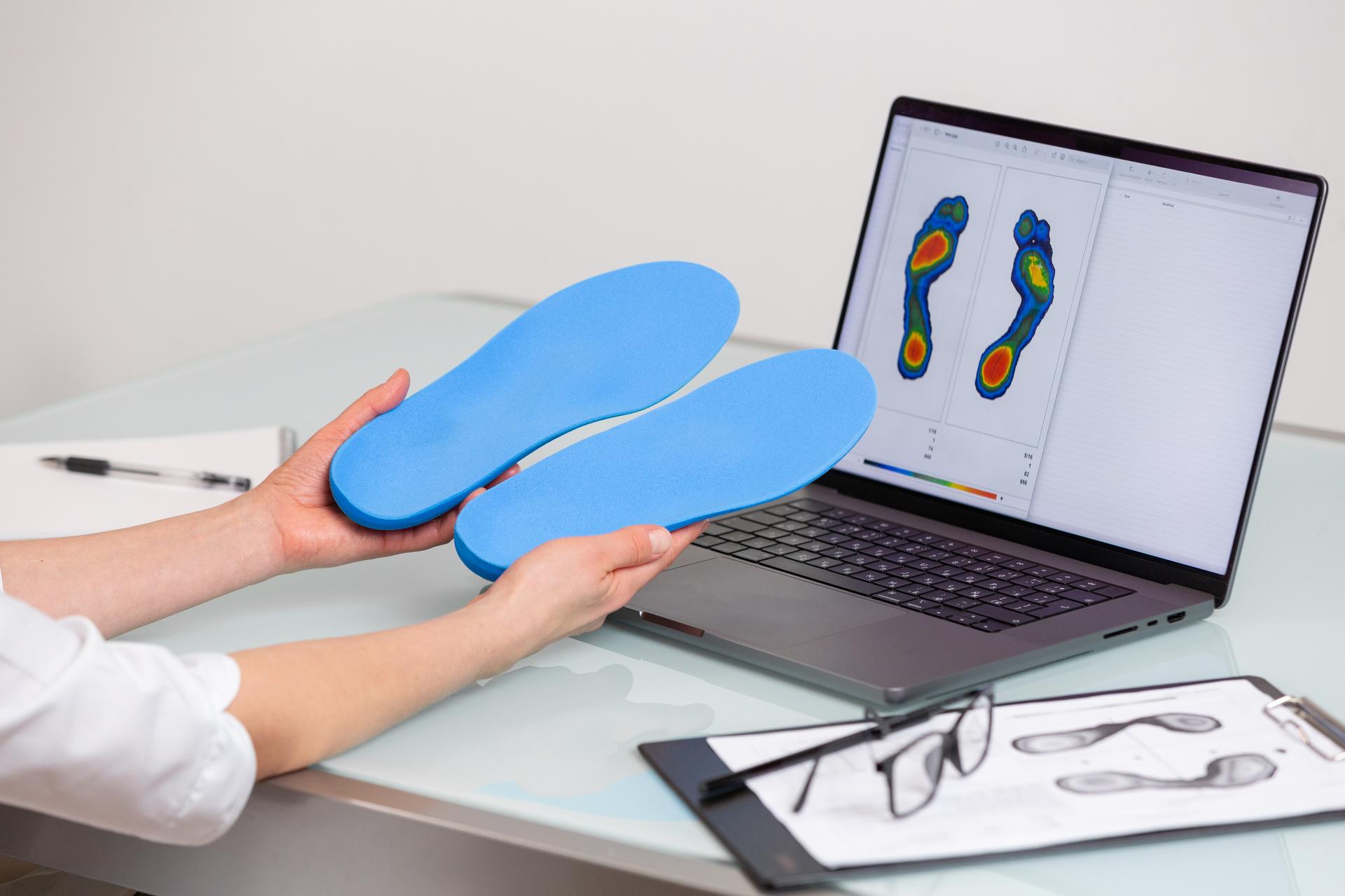 Female doctor orthopedist  holding custom made insoles in a clinic in front of the laptop with a test feet picture.  Feet recreation and orthotic medicine concept